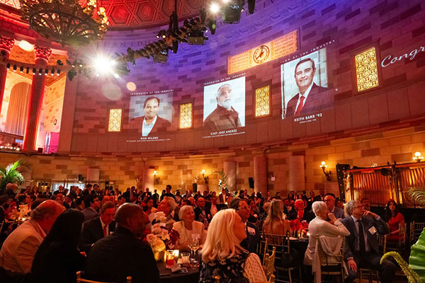 Photo of an elegant restaurant with a high ceiling and a huge chandelier hanging high above all. The room is filled with people sitting at tables and images of the evening’s honorees are projected onto the stone walls above the room’s columns.